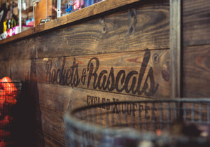 A rustic wooden counter with the words 'Rockets & Rascals' and 'CYCLES X COFFEE' printed on it, photographed in a real indoor setting with shallow depth of field.