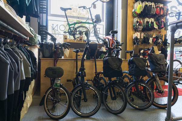 A row of Brompton bicycles with rear bags displayed inside Rockets and Rascals, Poole. Clothing racks, wall-mounted accessories, and a window are visible in the background.