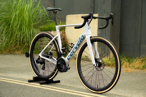 A custom built white Cervelo road bicycle with black components and tan sidewall tires, displayed on a black stand on an outdoor paved surface. Built by Rockets and Rascals in Poole, Dorset.