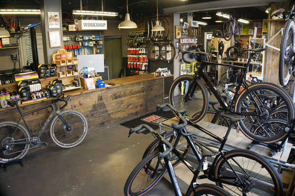 The Rockets and Rascals shop interior in Poole with multiple road and gravel bikes displayed on racks and the floor. A wooden counter with tools and supplies sits on the left, and shelves and signage are visible throughout the space.