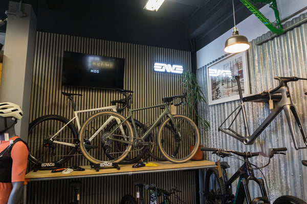 A showroom interior featuring multiple bicycles displayed on racks and stands against slatted wood and corrugated metal walls. A wall-mounted screen and illuminated ENVE logos are visible, along with a hanging pendant light and a framed print.