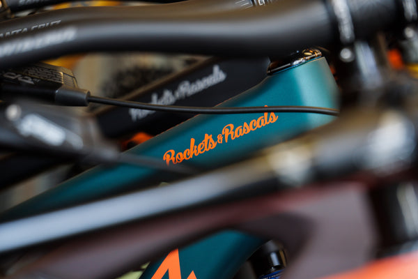 Close-up of a Santa Cruz bicycle frame in matte teal with orange script reading 'Rockets of Rascals' on the top tube, surrounded by blurred black handlebars and components.