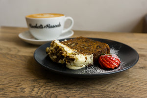 A slice of layered carrot cake on a black plate, served with a strawberry and dusted with powdered sugar, on a wooden table next to a flat white coffee made with Bad Hand coffee at Rocket's and Rascals bicycle cafe, Poole.